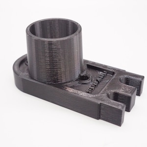 Puede incluir: Una pieza de plástico negra impresa en 3D con una abertura cilíndrica y una base rectangular. La base tiene una ranura en un lado y un pequeño agujero en el centro.