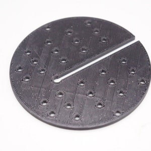 May include: A black circular 3D printed plate with a slit in the middle and a grid of small holes.