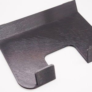 May include: A dark brown, wooden phone stand with a unique design. The stand features a rectangular base with a curved cutout and two small, rectangular hooks. The wood grain is visible.