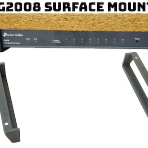 May include: A black surface mount bracket for a network device. The bracket is made of plastic and has two holes for mounting screws. The bracket is designed to be mounted to a surface, such as a desk or wall, and then the network device can be mounted to the bracket.