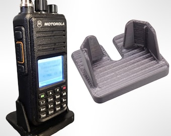 Pode incluir: Um rádio bidirecional Motorola preto com um visor digital e um teclado. O rádio está sentado em um suporte de plástico preto. Um suporte cinza impresso em 3D também é mostrado na imagem.