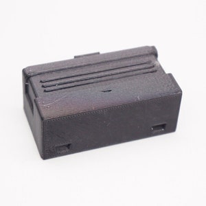 May include: A small, black, rectangular box with a textured surface and a series of raised lines on the top. The box has two small rectangular openings on the bottom.