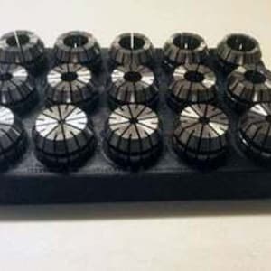 Puede incluir: Una bandeja de plástico negro con 16 mandriles de collet de metal plateado. Los mandriles están dispuestos en cuatro filas de cuatro.