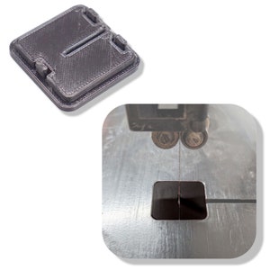 May include: A close-up of a black plastic jig being cut by a band saw. The jig is square and has a slot in the center. The band saw blade is visible in the center of the jig.
