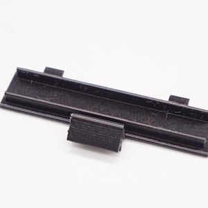 May include: A black plastic 3D printed part with a long rectangular shape and a smaller rectangular piece attached to one side.
