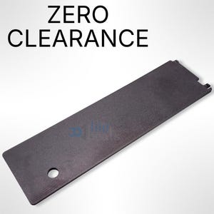 Puede incluir: Una pieza de plástico negra rectangular con un agujero en el centro. El texto "ZERO CLEARANCE" está impreso en blanco sobre un fondo blanco.
