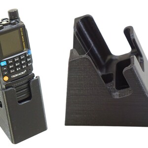 May include: A black handheld radio with a screen and keypad is in a black plastic charging stand. The stand is angled and has a textured surface. The radio has the word "TIDRADIO" printed on it.