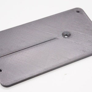 May include: A gray plastic rectangular plate with four holes and a slot in the center. The plate is designed for a 3D printer.
