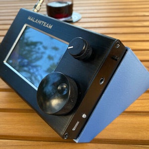 Puede incluir: Un dispositivo electrónico negro con una pequeña pantalla, dos botones y un soporte azul. El dispositivo tiene el texto "MALAMTEAM" impreso en la parte delantera.