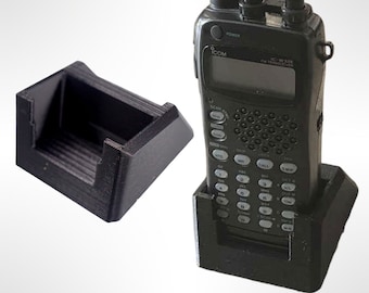 Pode incluir: Um suporte de carregamento preto para um rádio portátil com um rádio preto sentado no suporte. O rádio tem uma tela que diz "IC-W32E FM TRANSCEIVER" e um teclado com letras e números.