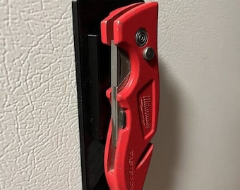 May include: A red Milwaukee Fastback utility knife with a black plastic handle is attached to a black magnetic wall mount.