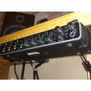 May include: A black audio interface with multiple input jacks, knobs, and buttons. The device is mounted under a wooden shelf and has a power cable connected. The interface is designed for audio recording and mixing.