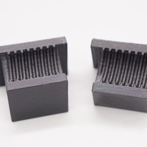 Puede incluir: Dos objetos de plástico negro impresos en 3D con una superficie texturizada. Los objetos tienen la forma de un pequeño cubo con una sección elevada en un lado.