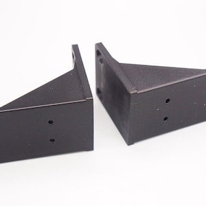 Könnte beinhalten: Zwei schwarze Kunststoffhalterungen mit mehreren Löchern. Die Halterungen sind abgewinkelt und haben auf einer Seite eine flache Oberfläche.