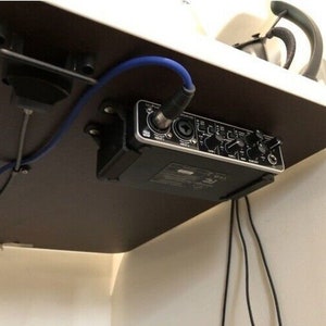 Könnte beinhalten: Ein schwarzes Audio-Interface mit Knöpfen und Reglern ist unter einem Schreibtisch mit schwarzen und blauen Kabeln montiert.