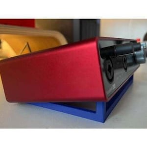 May include: A red audio interface with two input jacks and a blue stand. The audio interface is on a white surface.