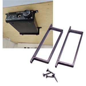 May include: Two black metal brackets with screws for mounting a device under a desk or shelf. The brackets are designed to hold a device with a rectangular shape.