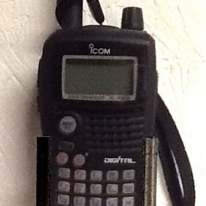 Puede incluir: Una radio bidireccional digital Icom negra con una pantalla pequeña y un teclado. La radio tiene una antena negra y una correa negra.