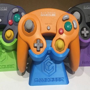 Puede incluir: Tres mandos de Nintendo GameCube en diferentes colores: morado, naranja y azul, con el logotipo de Nintendo GameCube en cada uno. Los mandos se muestran en soportes del mismo color.