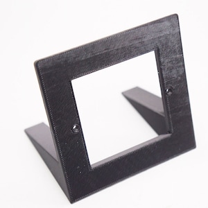 Könnte beinhalten: Ein schwarzer Wandplattenständer aus Kunststoff mit einer quadratischen Öffnung in der Mitte. Der Ständer hat zwei abgewinkelte Beine zur Unterstützung.