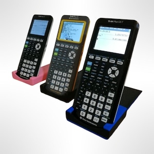 May include: Three graphing calculators with black cases and colour screens are displayed on stands. The stands are pink, black, and blue. The calculator on the right has the text "TI-84 Plus CE-T" on the top of the case.