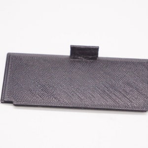 May include: A black plastic battery cover with a textured surface.