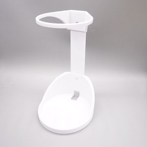May include: A white plastic stand with a circular base and a vertical support. The stand has a curved top ring and a slot in the base. The design is simple and functional, suitable for holding an item.