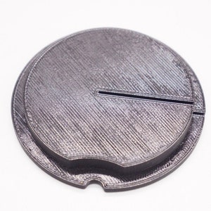 May include: A black 3D printed circular object with a groove around the edge and a small slit on the side.