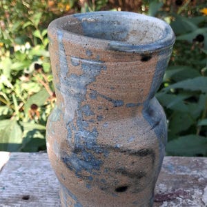 May include: A hand-thrown ceramic vase with a bulbous body and a flared rim. The vessel features a textured, tan-colored clay base with irregular, mottled blue glaze accents applied in vertical streaks and patches across the exterior surface.