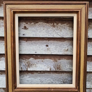 May include: A rectangular, empty picture frame with a gold and brown finish. The frame has a layered design with a lighter inner border and a darker outer edge. The background is a weathered wooden surface.