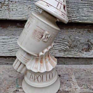 May include: A cream-colored ceramic decanter shaped like a space capsule with brown painted details. The base features the word APOLLO in brown lettering. The design includes a conical top, a cylindrical midsection, and a flared, fluted base.