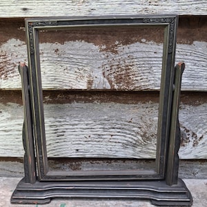 May include: An antique, rectangular picture frame with a dark, distressed finish. The frame is supported by two vertical posts and a base, all in a matching dark color. The frame has decorative detailing on the corners and along the top and bottom edges.