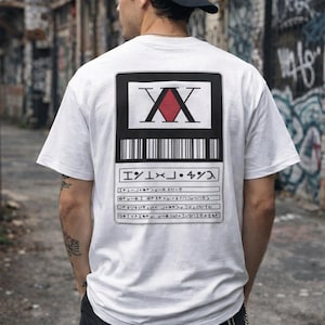 Puede incluir: Camiseta blanca con un gráfico en la espalda que presenta un rectángulo negro con las letras "XX" y un rombo rojo. Debajo del gráfico hay caracteres japoneses y un código de barras. La persona lleva una gorra de béisbol negra.