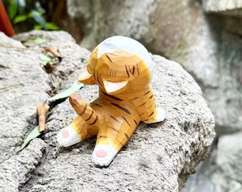 May include: A small hand-carved wooden figurine of a tabby cat with orange fur and dark brown stripes. The cat is depicted in a seated position, facing away from the viewer, with a white patch on its head, white paws, and small pink paw pads.