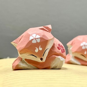 May include: Two pink wooden fox figurines with white flower designs and pink accents. The foxes are curled up in a sleeping position.
