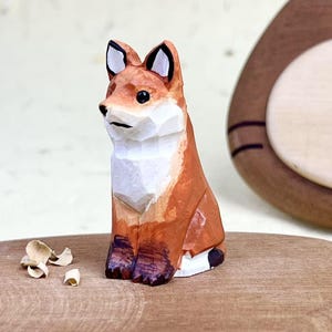 May include: A small, hand-carved wooden fox figurine featuring faceted geometric surfaces. The fox is painted in shades of orange, white, and dark brown, with black accents on the ears and eyes, showcasing a distinct carved texture throughout.