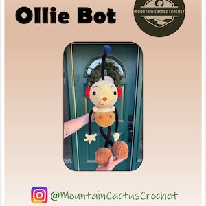 May include: A crocheted amigurumi toy called Ollie Bot. The toy is brown, beige, and black with a blue antenna. The toy is being held in front of a green door. The image includes the text "Ollie Bot" and "Mountain Cactus Crochet".