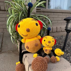 May include: Two crocheted plush toys shaped like bees. The larger bee is yellow with brown legs and a black antenna. The smaller bee is yellow with brown legs and a red antenna. Both bees have black eyes and a smiling face.