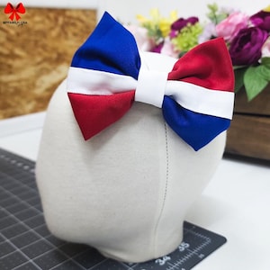 May include: A red, white, and blue bow made of fabric. The bow is tied in a knot and has a white stripe in the middle.