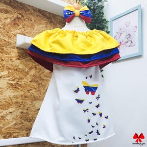 May include: A white dress with a yellow, blue and red ruffled top and a pattern of butterflies with the Venezuelan flag on their wings.