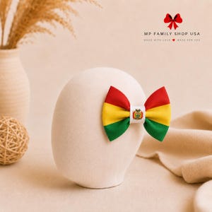 Puede incluir: Un lazo para el cabello con la bandera tricolor horizontal de Bolivia en rojo, amarillo y verde. El centro del lazo está sujeto con una banda blanca que muestra el escudo de armas boliviano. El texto MP FAMILY SHOP USA es visible arriba.