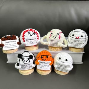 Puede incluir: Una colección de personajes deportivos hechos a mano, tejidos a ganchillo, cada uno con un diseño único y un pequeño letrero con un mensaje inspirador. Los personajes incluyen un balón de fútbol americano, un balón de béisbol, un balón de baloncesto y una pelota de golf.