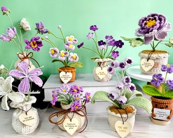 May include: A collection of handmade crochet flowers in various shades of purple, white, and yellow, displayed in small, decorative pots. Each pot has a unique design and a wooden heart-shaped tag with text. The flowers include lilies, pansies, and other floral designs.