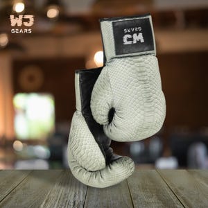 Puede incluir: Un par de guantes de boxeo con un patrón de piel de serpiente en verde claro y negro. Los guantes tienen el texto "W3 GEARS" y "CM". Los guantes se muestran sobre una superficie de madera.