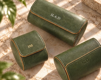 May include: Three dark green cylindrical watch cases with rounded edges and contrasting tan trim. Each case features a flap closure and is personalised with gold embossed initials: H.A.D on the largest, JM on the smallest, and K.D on the other.
