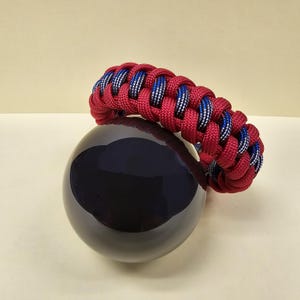 May include: A red and blue braided paracord bracelet. The bracelet features a repeating pattern of red, blue, and white strands. The bracelet is resting on a dark blue, spherical object.