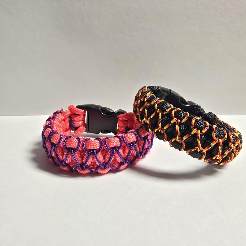 Pu&ograve; includere: Due braccialetti in paracord con fibbie in plastica nera. Uno &egrave; rosa corallo con dettagli viola, l'altro &egrave; nero con dettagli gialli e arancioni. Entrambi sono su una superficie bianca.