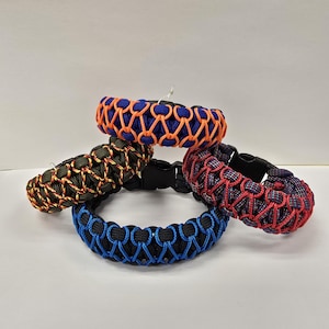 Pu&ograve; includere: Quattro braccialetti in paracord di vari colori: blu con arancione, verde con arancione, rosso con blu e nero con blu. Ogni braccialetto presenta un design intrecciato e una chiusura in plastica nera. I braccialetti sono esposti su una superficie bianca.
