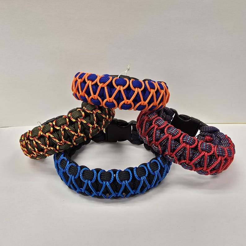 Pu&ograve; includere: Quattro braccialetti in paracord di vari colori: blu con arancione, verde con arancione, rosso con blu e nero con blu. Ogni braccialetto presenta un design intrecciato e una chiusura in plastica nera. I braccialetti sono esposti su una superficie bianca.