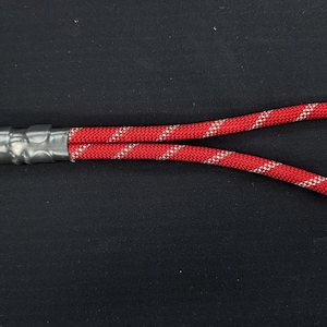Può includere: Un cinturino da polso in corda rossa con un motivo diagonale argento riflettente su tutto il cordino. La corda è ripiegata ad anello e fissata alla base con un nastro adesivo grigio, collegato a un moschettone girevole in metallo argentato.